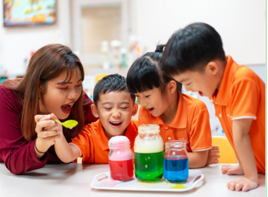 A My First Skool teacher in charge of a small group of three smiling preschool children, so as to build feelings of trust and safety as part of the relationship-based curriculum